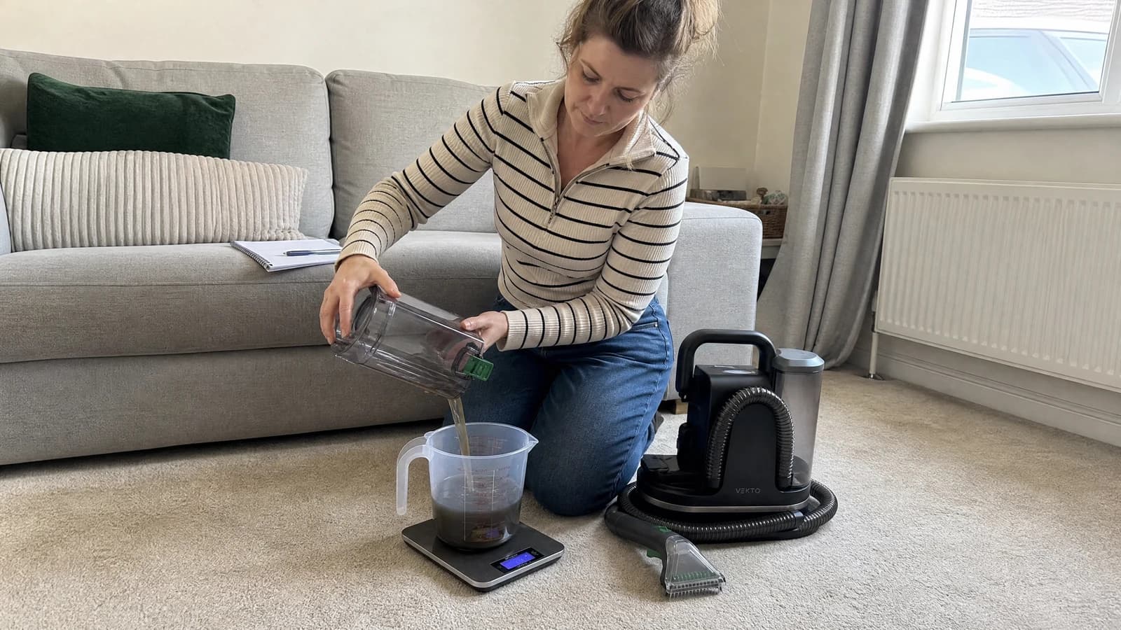 A tester kneeling by a grey fabric sofa, pouring a dirty tank into a measuring jug on a kitchen scale. The Vekto Spot Cleaner P30 on the carpet beside her