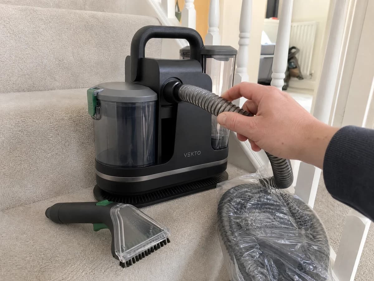 A hand unplugging the hose from the Vekto Spot Cleaner P30 body on a cream stair carpet, the spare hose in its bag beside it