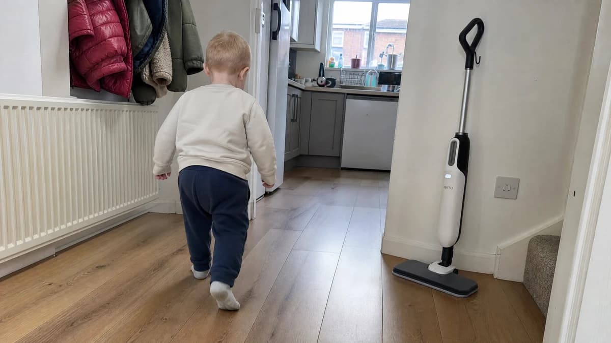 A toddler in socks walking across a dry laminate hallway towards the kitchen, the Vekto Steam Mop S20 standing by the wall with its light on