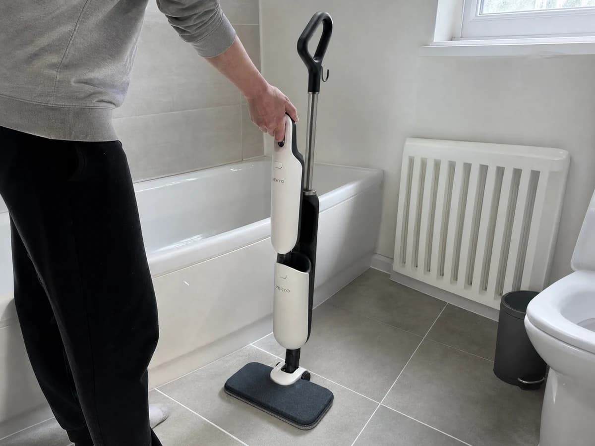 A hand lifting the steam unit out of the Vekto Steam Mop S20 body in a tiled bathroom, with a grey grout line on the floor beside it