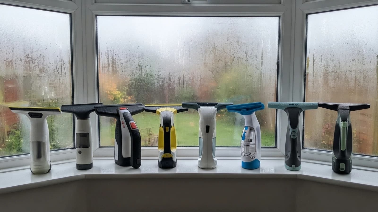 Eight cordless window vacs standing in a row on a uPVC window sill in a bay window fogged with condensation, the Vekto Window Vac W10 second from left