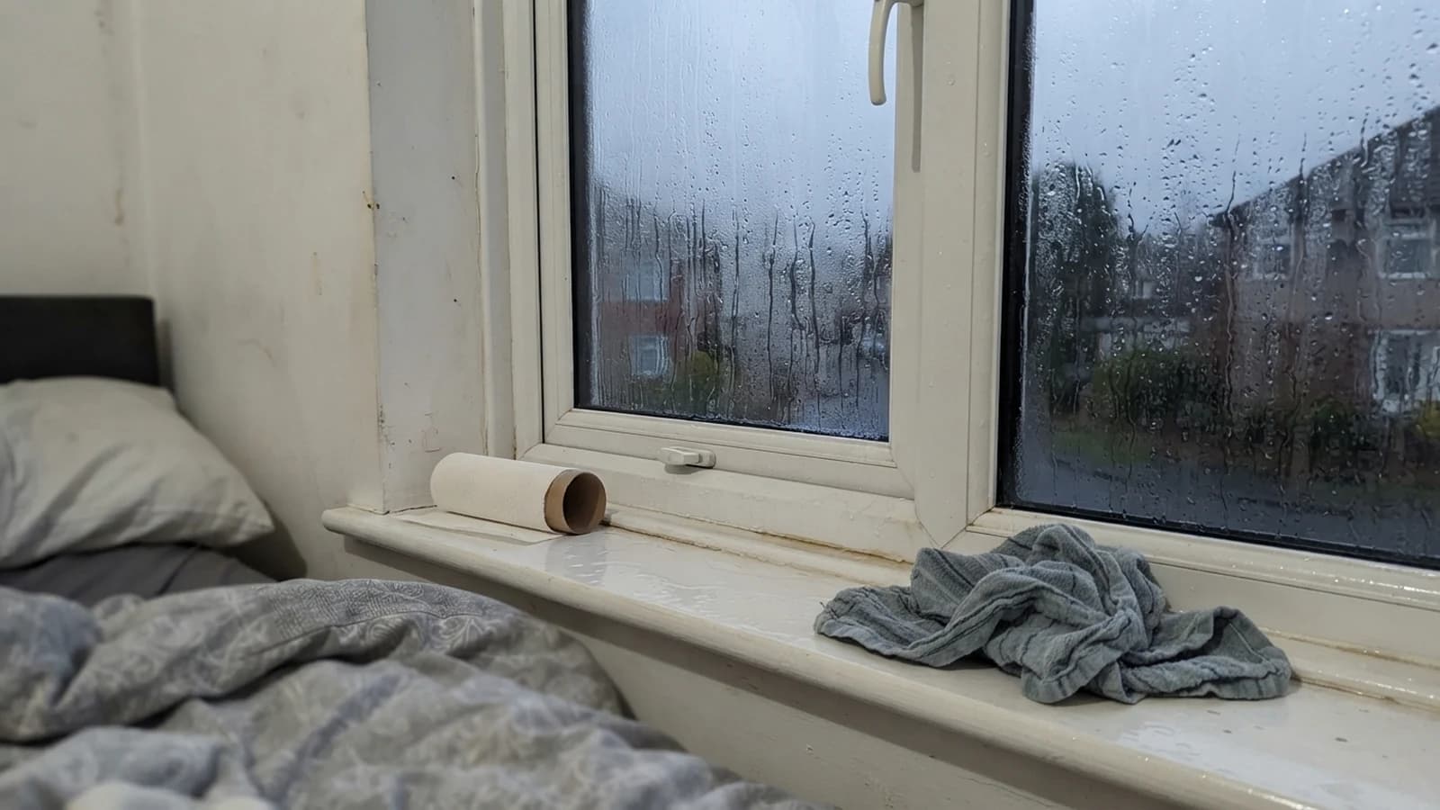 A bedroom uPVC window streaming with condensation at dawn, water pooled along the bottom seal, a roll of kitchen paper and a damp tea towel on the sill