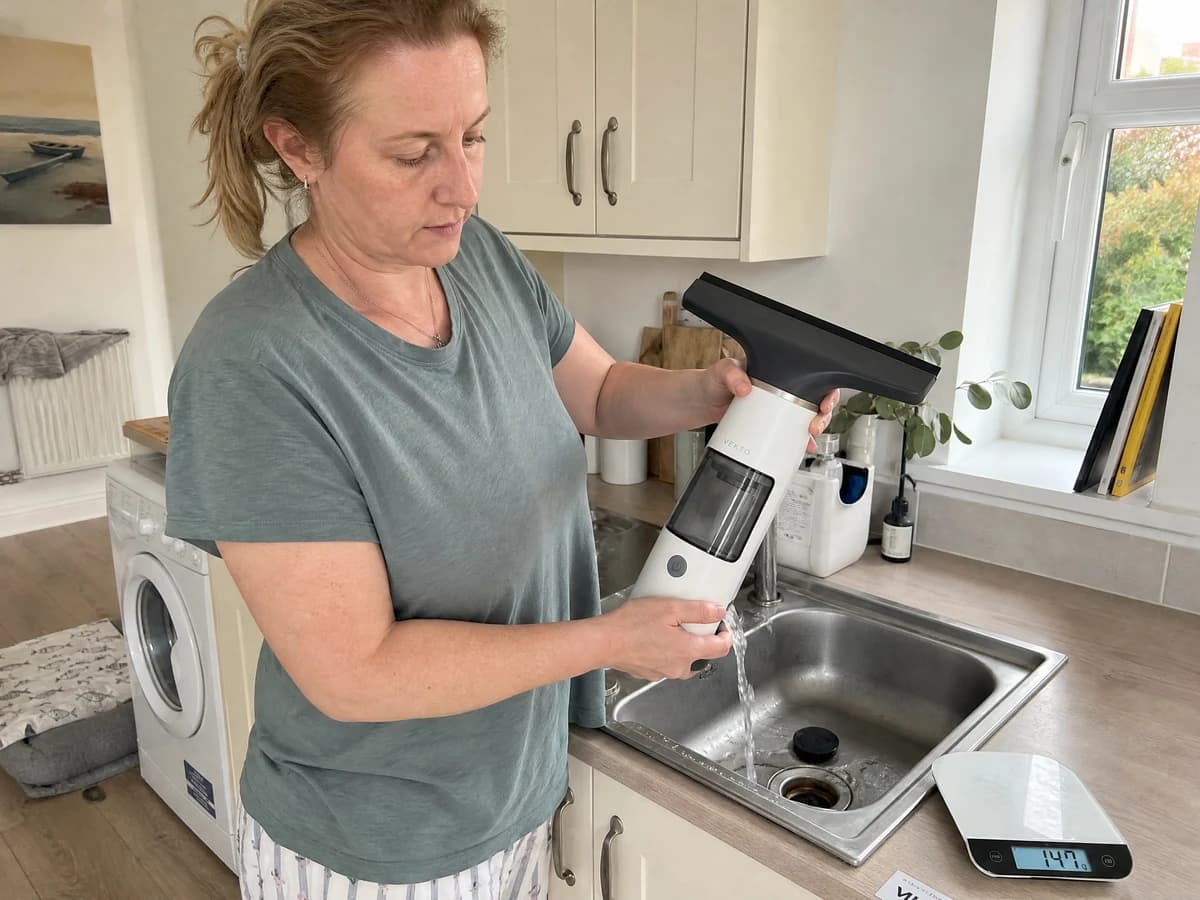 The Vekto Window Vac W10 tank held over a kitchen sink with its drain cap open and grey water pouring out, a kitchen scale beside it reading 147 g