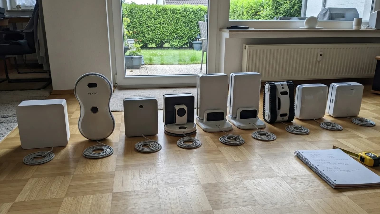 Neun Fensterputzroboter stehen in einer Reihe auf einem Holzboden vor einer bodentiefen Terrassentür, der Vekto Fensterputzroboter als zweiter von links