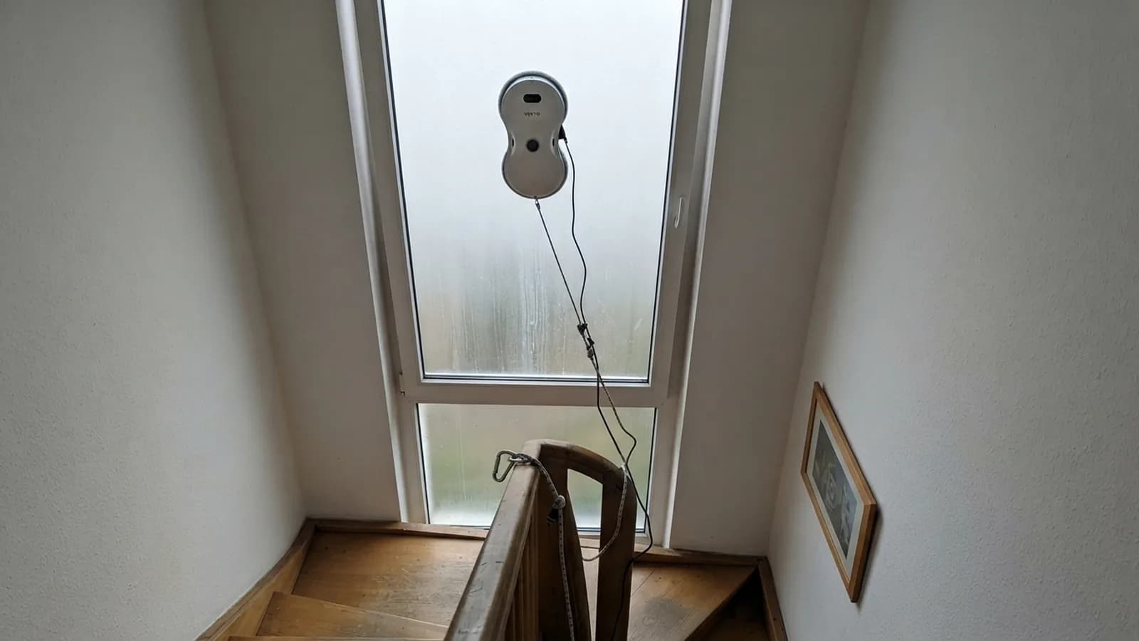 Der Vekto Fensterputzroboter putzt oben ein hohes Fenster im Treppenhaus, das Seil hängt nach unten