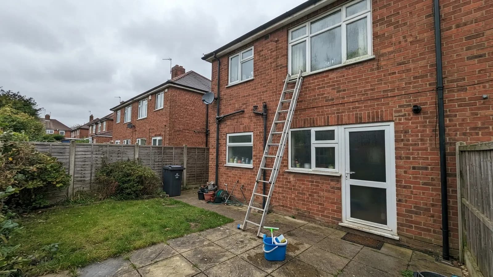 An aluminium ladder leaning against the back of a red-brick semi under a first-floor window, a bucket with a squeegee on the patio below