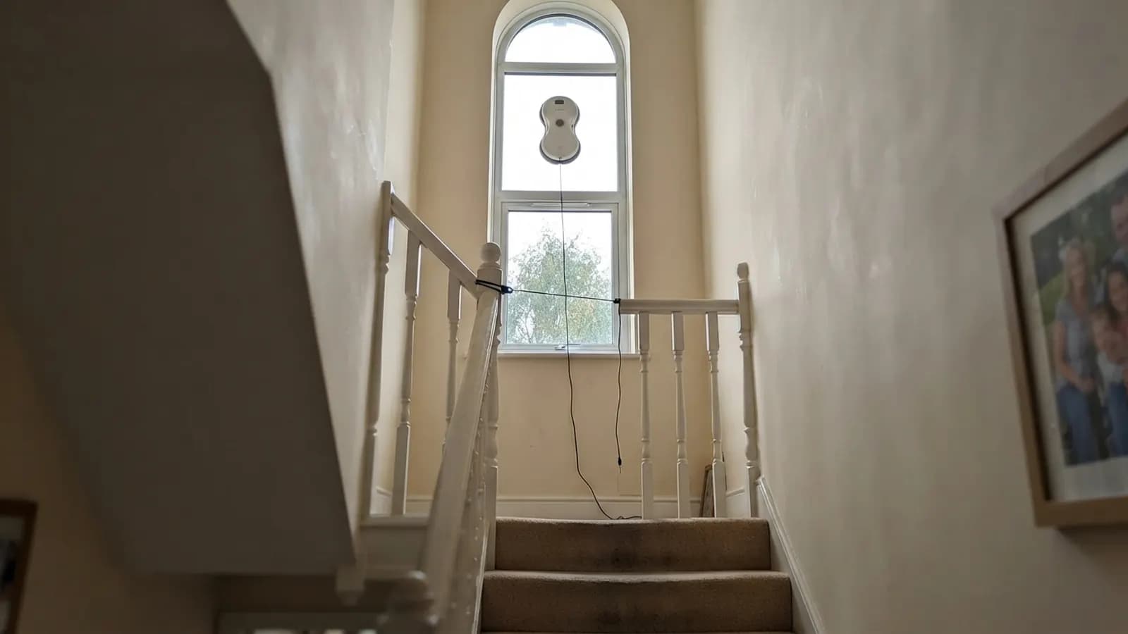 The Vekto Window Robot cleaning a tall arched landing window high above a carpeted staircase, its rope clipped to the banister