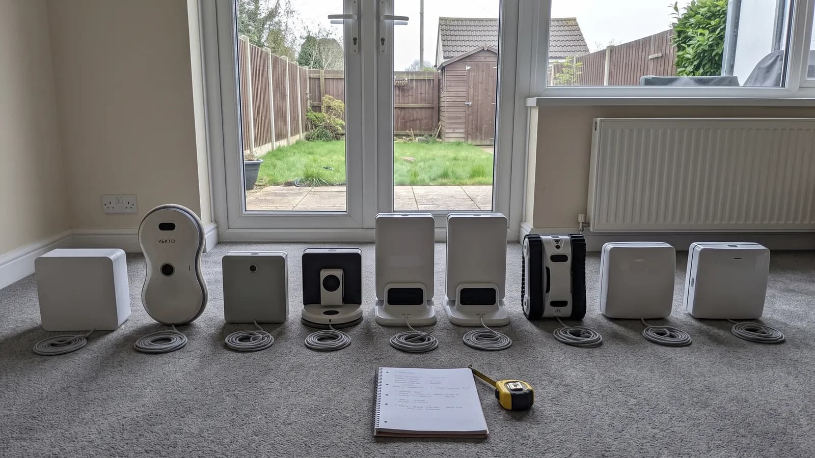 Nine window cleaning robots in a row on a living room floor in front of white uPVC patio doors, the Vekto Window Robot second from left