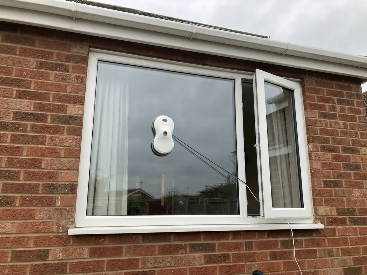 The Vekto Window Robot cleaning the outside of a white uPVC window in a red-brick wall, its safety rope running in through the open casement next to it