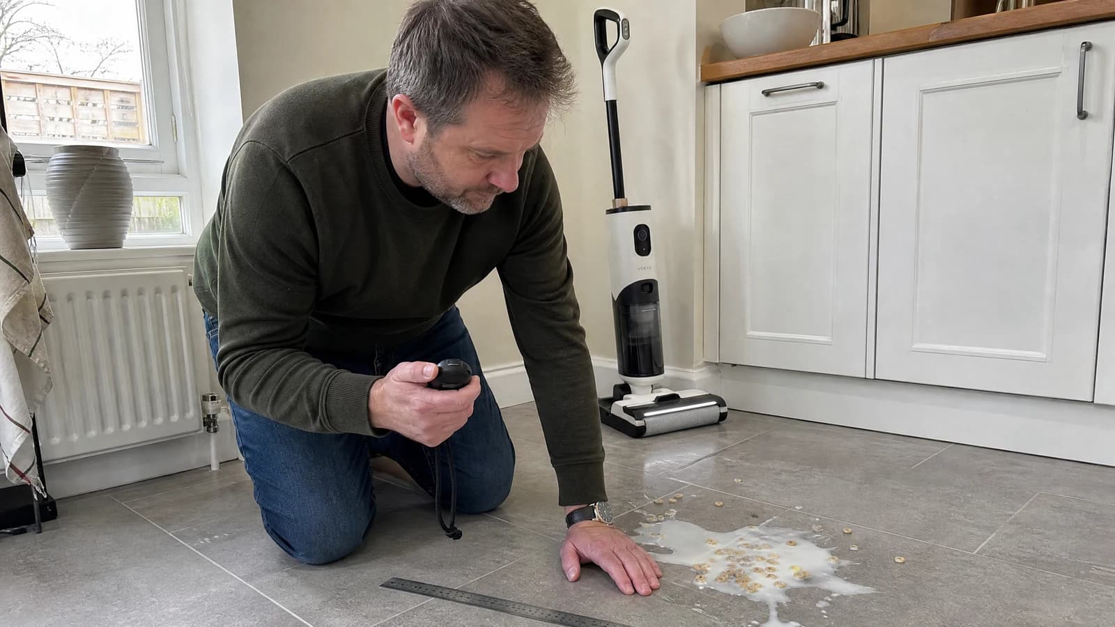 A tester kneeling on a tiled kitchen floor with a stopwatch and a steel ruler beside a spill of milk and cereal, the Vekto Floor Washer F40 standing behind