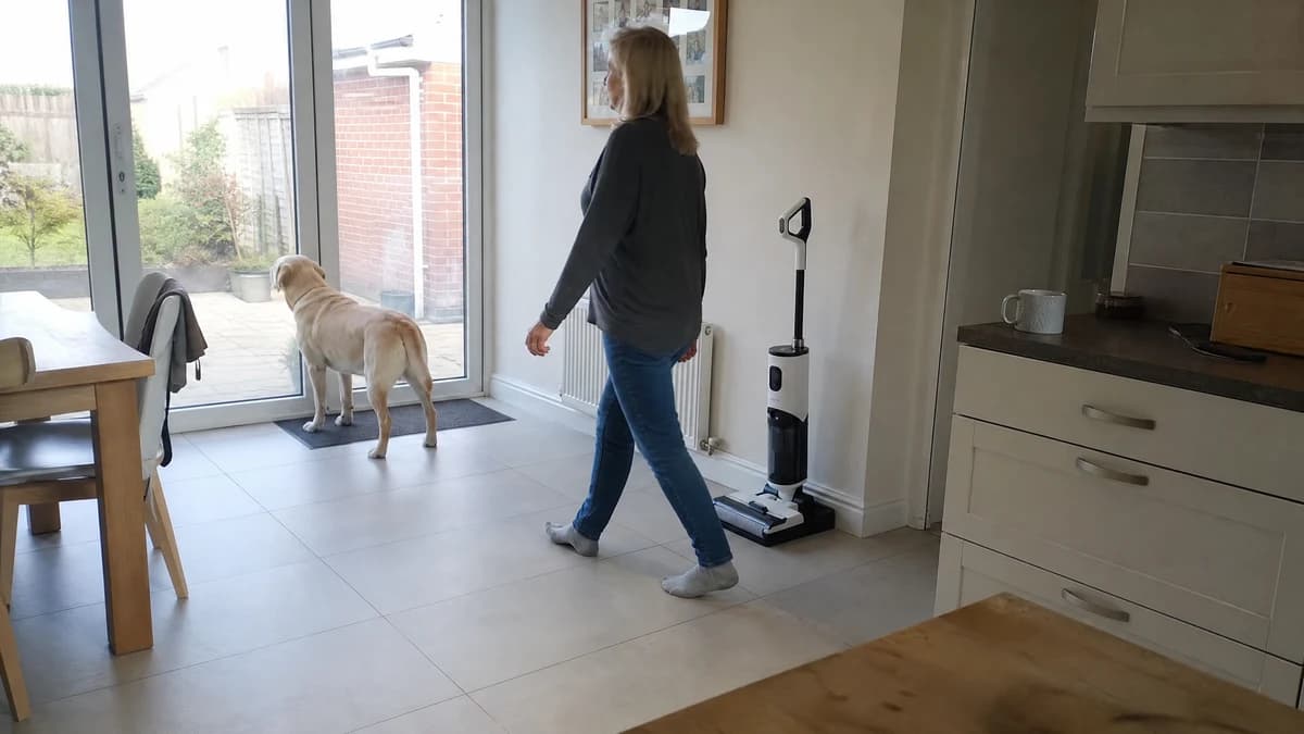 A woman in socks walking across a dry tiled kitchen-diner towards a labrador at the patio door, the Vekto Floor Washer F40 standing on its dock in the corner