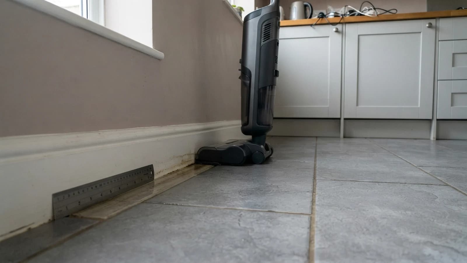 A cordless floor washer standing in the corner of a tiled kitchen-diner with a ruler on the floor showing a dirty strip left along the skirting board