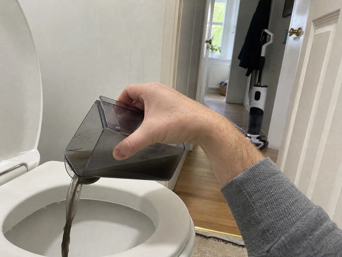 A hand tipping the Vekto Floor Washer F40 dirty tank of black water into a toilet, the washer standing on its dock in the background