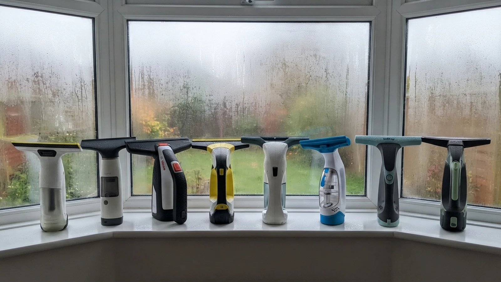 Eight cordless window vacs standing in a row on a uPVC window sill in a bay window fogged with condensation, the AIRVAC Window Vac W10 second from left